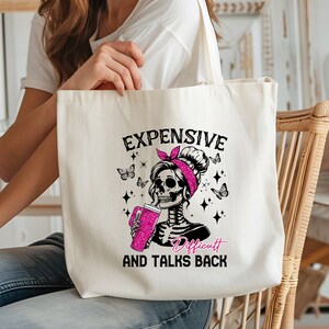 Op de afbeelding: Cr&egrave;mekleurige draagtas met een skeletafbeelding met een roze haarband en een roze drankje. De tekst op de tas luidt "EXPENSIVE, DIFFICULT, AND TALKS BACK". Vlinders en sterren zijn ook afgebeeld.