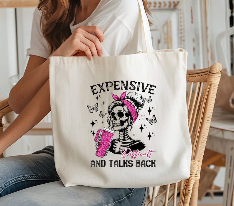 Op de afbeelding: Cr&egrave;mekleurige draagtas met een skeletafbeelding met een roze haarband en een roze drankje. De tekst op de tas luidt "EXPENSIVE, DIFFICULT, AND TALKS BACK". Vlinders en sterren zijn ook afgebeeld.