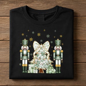 May include: A black t-shirt featuring a graphic print of two green nutcrackers standing on either side of a Christmas tree decorated with a large bow and surrounded by wrapped gifts, all accented with gold glitter and small snowflake icons.