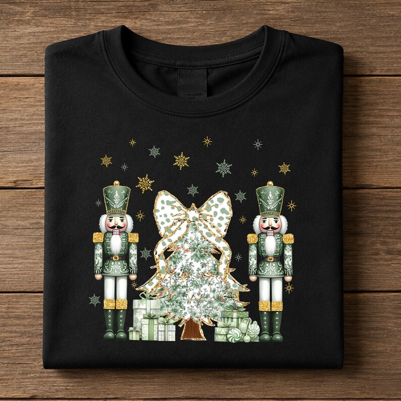 May include: A black t-shirt featuring a graphic print of two green nutcrackers standing on either side of a Christmas tree decorated with a large bow and surrounded by wrapped gifts, all accented with gold glitter and small snowflake icons.