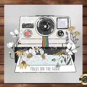 May include: A vintage-style instant camera with a rainbow film strip and a white frame with the text "Focus on the good". The camera is surrounded by white and yellow flowers.