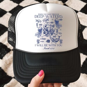 May include: Black and white trucker hat with a white front panel featuring a blue illustration of sea creatures and the text "WHEN YOU PASS THROUGH DEEP WATERS, I WILL BE WITH YOU, Isaiah 43:2". The hat has a mesh back.