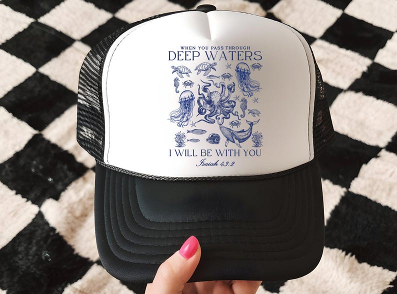 May include: Black and white trucker hat with a white front panel featuring a blue illustration of sea creatures and the text "WHEN YOU PASS THROUGH DEEP WATERS, I WILL BE WITH YOU, Isaiah 43:2". The hat has a mesh back.