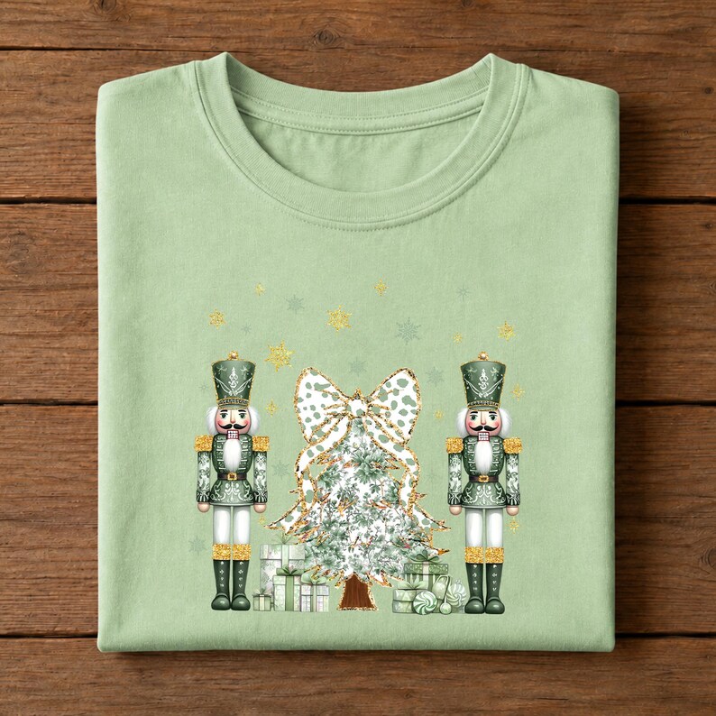 May include: A light sage green t-shirt featuring a graphic print of two nutcrackers standing on either side of a decorated Christmas tree. The tree is topped with a large white and green patterned bow, and small gold stars are scattered above.