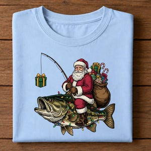 May include: A light blue t-shirt featuring a graphic of Santa Claus riding a large green bass fish. Santa holds a fishing rod with a gift box as bait, and a sack of presents is strapped to his back. The fish is decorated with colorful Christmas lights.