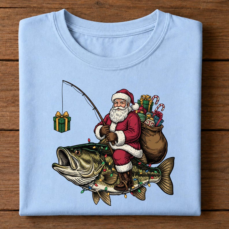 May include: A light blue t-shirt featuring a graphic of Santa Claus riding a large green bass fish. Santa holds a fishing rod with a gift box as bait, and a sack of presents is strapped to his back. The fish is decorated with colorful Christmas lights.