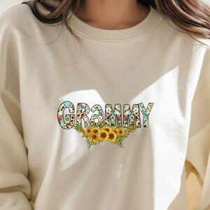 Op de afbeelding: Een cr&egrave;mekleurig sweatshirt met het woord "GRAMMY" bedrukt in kleurrijke letters met een bloemen- en luipaardprint. De letters worden omringd door een bloemendesign met zonnebloemen.