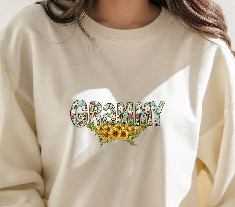 Op de afbeelding: Een cr&egrave;mekleurig sweatshirt met het woord "GRAMMY" bedrukt in kleurrijke letters met een bloemen- en luipaardprint. De letters worden omringd door een bloemendesign met zonnebloemen.