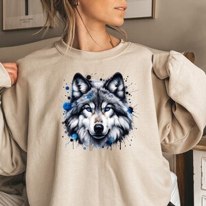 May include: A beige sweatshirt with a watercolor-style wolf graphic. The wolf has blue eyes and is surrounded by blue and black paint splatters.