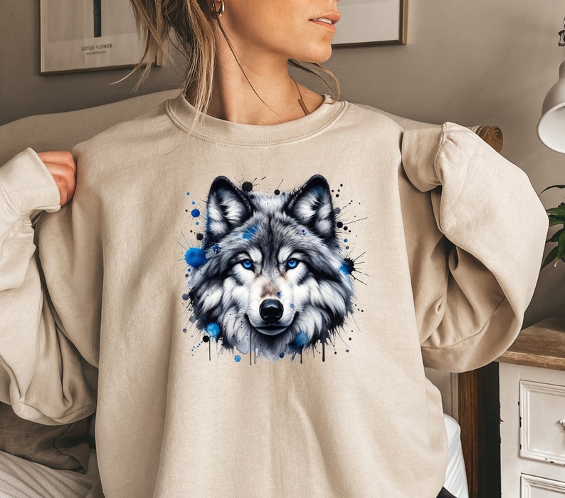 May include: A beige sweatshirt with a watercolor-style wolf graphic. The wolf has blue eyes and is surrounded by blue and black paint splatters.