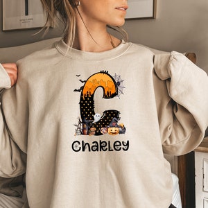 May include: Beige sweatshirt featuring a Halloween-themed design. The design includes a large letter "C" filled with orange and black elements, a ghost, a witch's hat, and a jack-o'-lantern. The name "Chakley" is printed below the letter in black.