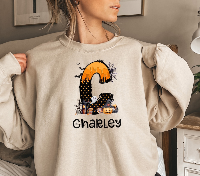 May include: Beige sweatshirt featuring a Halloween-themed design. The design includes a large letter "C" filled with orange and black elements, a ghost, a witch's hat, and a jack-o'-lantern. The name "Chakley" is printed below the letter in black.