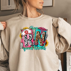 May include: A beige sweatshirt with a graphic design of the letters "RN" in a colorful, patterned style. The letters are surrounded by medical symbols and the text "You good hands".