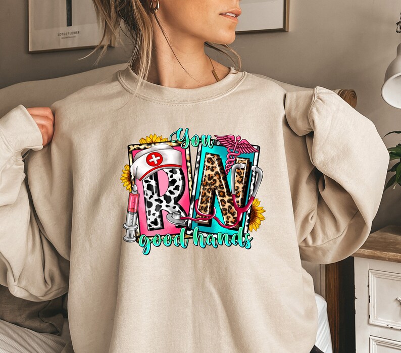 May include: A beige sweatshirt with a graphic design of the letters "RN" in a colorful, patterned style. The letters are surrounded by medical symbols and the text "You good hands".