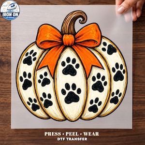 May include: A DTF iron-on transfer sheet featuring a white pumpkin illustration with black paw prints, a large orange bow, and a brown stem. The text at the bottom reads PRESS PEEL WEAR DTF TRANSFER, and the top left corner shows an iron icon with the text IRON ON.