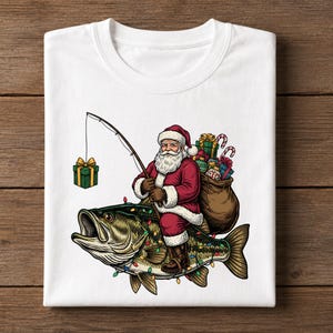 May include: A white t-shirt featuring a graphic of Santa Claus riding a large bass fish. Santa wears a red suit and carries a brown sack filled with gifts. He holds a fishing rod with a green gift box on the line. The fish is decorated with colorful Christmas lights.