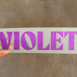 May include: A rectangular adhesive transfer sheet featuring the word VIOLET in large, bold, uppercase letters. The text has a distressed, weathered texture and a vibrant purple color, set against a clear, semi-transparent backing material.
