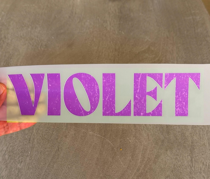 May include: A rectangular adhesive transfer sheet featuring the word VIOLET in large, bold, uppercase letters. The text has a distressed, weathered texture and a vibrant purple color, set against a clear, semi-transparent backing material.