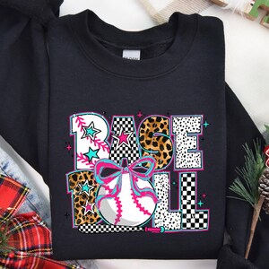 May include: Black sweatshirt with a graphic design of a baseball with a bow and the word "BASEBALL" in a colorful, patterned font.