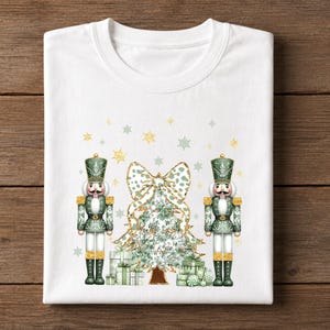 May include: A white crew neck t-shirt featuring a graphic print of two green and gold nutcrackers standing on either side of a decorated Christmas tree with a large leopard print bow on top, surrounded by small gold and green snowflakes.