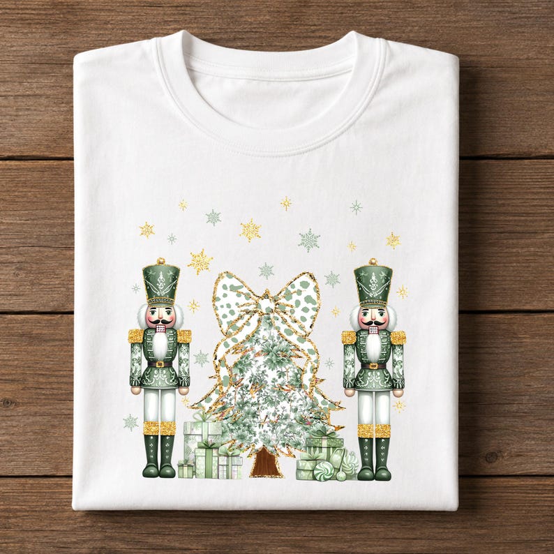 May include: A white crew neck t-shirt featuring a graphic print of two green and gold nutcrackers standing on either side of a decorated Christmas tree with a large leopard print bow on top, surrounded by small gold and green snowflakes.