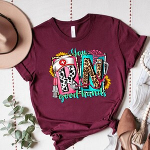 May include: A maroon t-shirt with a graphic design featuring the letters "RN" in a colorful, patterned style. The letters are surrounded by sunflowers, a stethoscope, a syringe, and a nurse's hat. The text "You good hands" is below the letters.