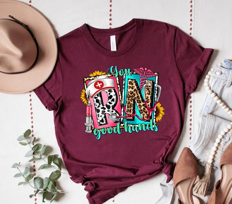 May include: A maroon t-shirt with a graphic design featuring the letters "RN" in a colorful, patterned style. The letters are surrounded by sunflowers, a stethoscope, a syringe, and a nurse's hat. The text "You good hands" is below the letters.