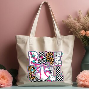 May include: A white canvas tote bag with a colorful graphic design that says "BASEBALL" with a baseball and bat. The graphic design includes a pink bow, leopard print, and checkered pattern.