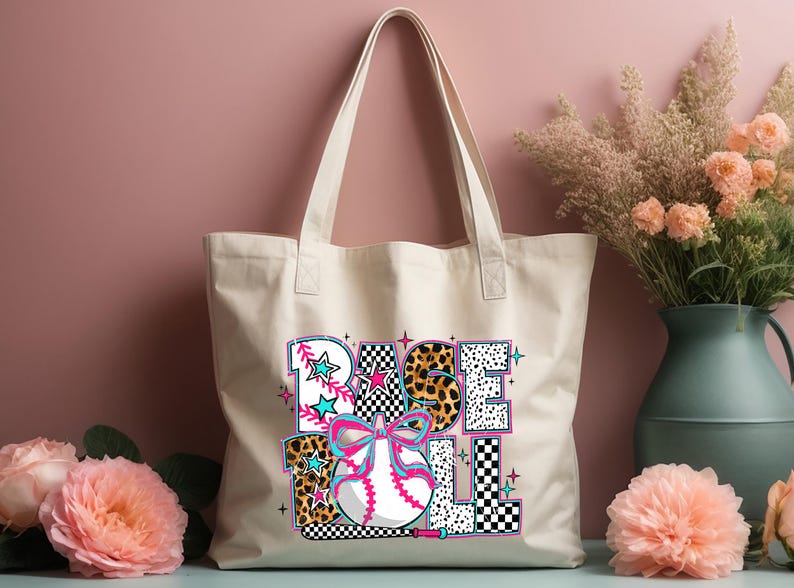 May include: A white canvas tote bag with a colorful graphic design that says "BASEBALL" with a baseball and bat. The graphic design includes a pink bow, leopard print, and checkered pattern.