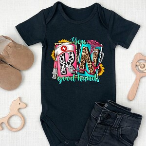 May include: Black baby onesie with a graphic print of the letters RN in a colorful design with a stethoscope, a syringe, a sunflower, and the text "You good hands".