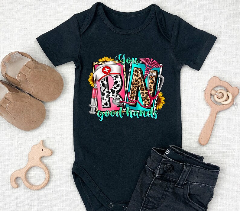 May include: Black baby onesie with a graphic print of the letters RN in a colorful design with a stethoscope, a syringe, a sunflower, and the text "You good hands".