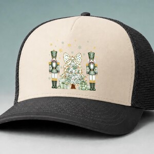 May include: A beige and black trucker hat featuring a graphic print of two green nutcrackers standing on either side of a decorated Christmas tree with a large bow on top and several wrapped gift boxes at the base, accented by gold stars.