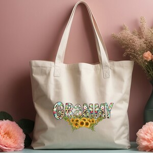 Op de afbeelding: Een witte canvas tote bag met het woord "GRAMMY" bedrukt in kleurrijke bloemenletters met een bloemen- en zonnebloemdessin.