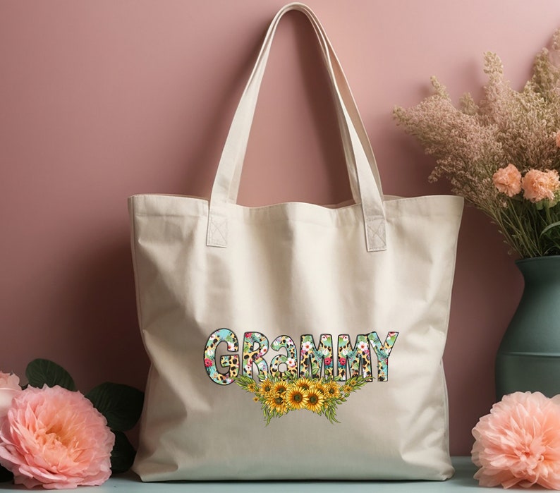 Op de afbeelding: Een witte canvas tote bag met het woord "GRAMMY" bedrukt in kleurrijke bloemenletters met een bloemen- en zonnebloemdessin.