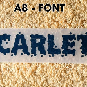 May include: A rectangular transfer sheet featuring the name SCARLETT in a dark blue, dripping, liquid-style font. The text is centered on a translucent backing, with the label A8 - FONT positioned above the design in a bold, black sans-serif typeface.