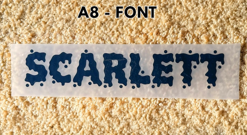 May include: A rectangular transfer sheet featuring the name SCARLETT in a dark blue, dripping, liquid-style font. The text is centered on a translucent backing, with the label A8 - FONT positioned above the design in a bold, black sans-serif typeface.