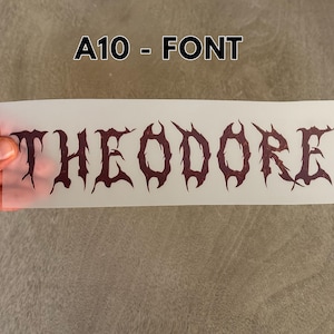 May include: A rectangular, semi-transparent transfer sheet featuring the name THEODORE printed in a dark, jagged, gothic-style font. The text is centered on the sheet, which is labeled A10 - FONT at the top. The letters have a distressed, textured appearance.