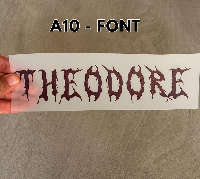 May include: A rectangular, semi-transparent transfer sheet featuring the name THEODORE printed in a dark, jagged, gothic-style font. The text is centered on the sheet, which is labeled A10 - FONT at the top. The letters have a distressed, textured appearance.
