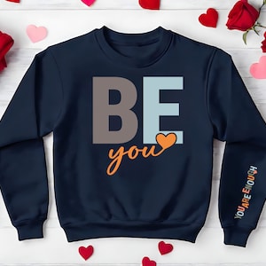 May include: Navy blue sweatshirt with the words "BE you" in orange, brown, and light blue lettering. A small orange heart is above the word "you". The right sleeve has the words "YOU ARE ENOUGH" in white lettering.