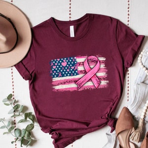 May include: A maroon t-shirt with a pink ribbon and an American flag design. The flag is distressed and has pink hearts and leopard print accents.