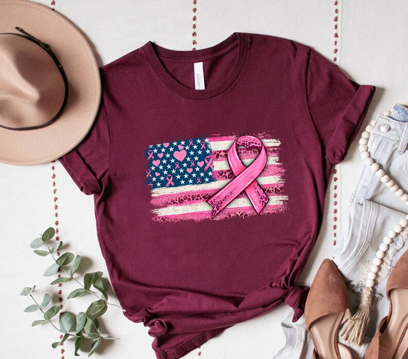 May include: A maroon t-shirt with a pink ribbon and an American flag design. The flag is distressed and has pink hearts and leopard print accents.