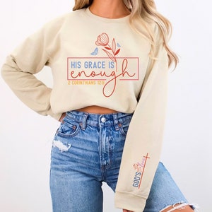 May include: A beige sweatshirt with a floral design and the text "HIS GRACE IS enough 2 CORINTHIANS 12:9" in red and blue. The sweatshirt has a red and blue cross with the text "GOD'S love" on the sleeve.