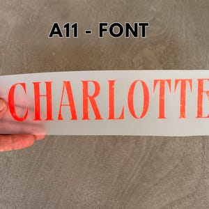 May include: A rectangular, semi-transparent transfer sheet featuring the name CHARLOTTE printed in a bold, serif, orange-red font with a distressed, textured appearance. The text A11 - FONT is displayed in black above the transfer sheet.