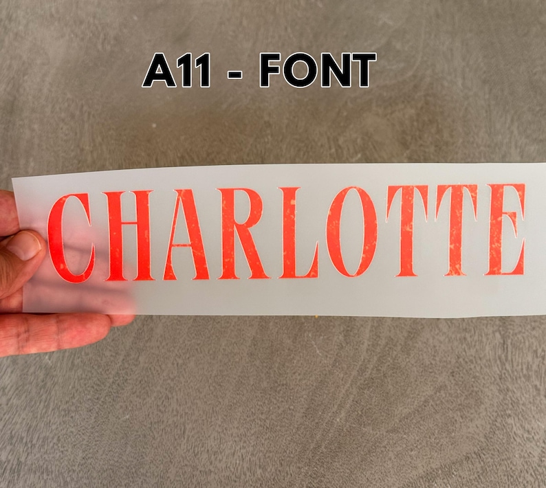 May include: A rectangular, semi-transparent transfer sheet featuring the name CHARLOTTE printed in a bold, serif, orange-red font with a distressed, textured appearance. The text A11 - FONT is displayed in black above the transfer sheet.