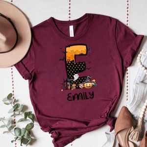 May include: Burgundy t-shirt featuring a Halloween-themed design. The design includes the letter "E" in orange and black, with a spooky house, pumpkin, and the name "Emily" in black. Bats and a spider are also included.