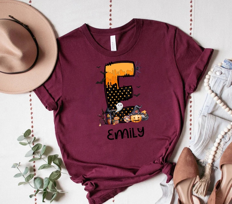 May include: Burgundy t-shirt featuring a Halloween-themed design. The design includes the letter "E" in orange and black, with a spooky house, pumpkin, and the name "Emily" in black. Bats and a spider are also included.