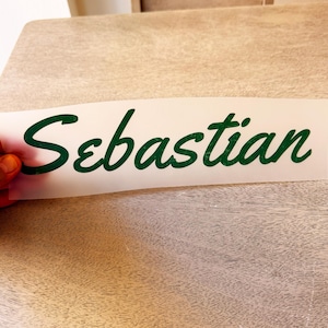May include: A rectangular white adhesive transfer sheet featuring the name Sebastian written in a dark green cursive script font. The lettering has a slightly textured appearance and is applied to a clear backing material for easy application.