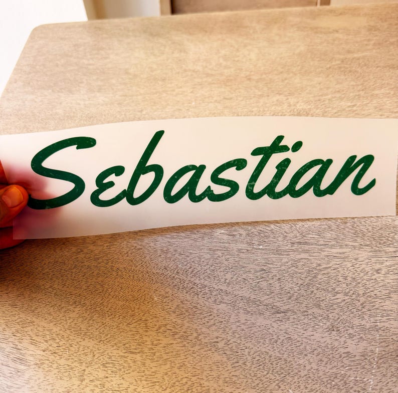 May include: A rectangular white adhesive transfer sheet featuring the name Sebastian written in a dark green cursive script font. The lettering has a slightly textured appearance and is applied to a clear backing material for easy application.