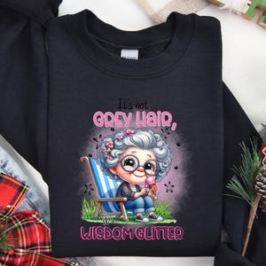 Op de afbeelding: Zwart sweatshirt met een cartoonillustratie van een vrouw met grijs haar en een bril die een ijsje vasthoudt. De tekst "It's not grey hair, it's wisdom glitter" staat op het sweatshirt gedrukt.