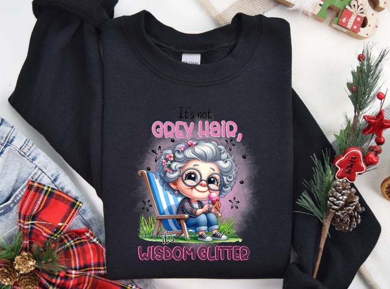 Op de afbeelding: Zwart sweatshirt met een cartoonillustratie van een vrouw met grijs haar en een bril die een ijsje vasthoudt. De tekst "It's not grey hair, it's wisdom glitter" staat op het sweatshirt gedrukt.